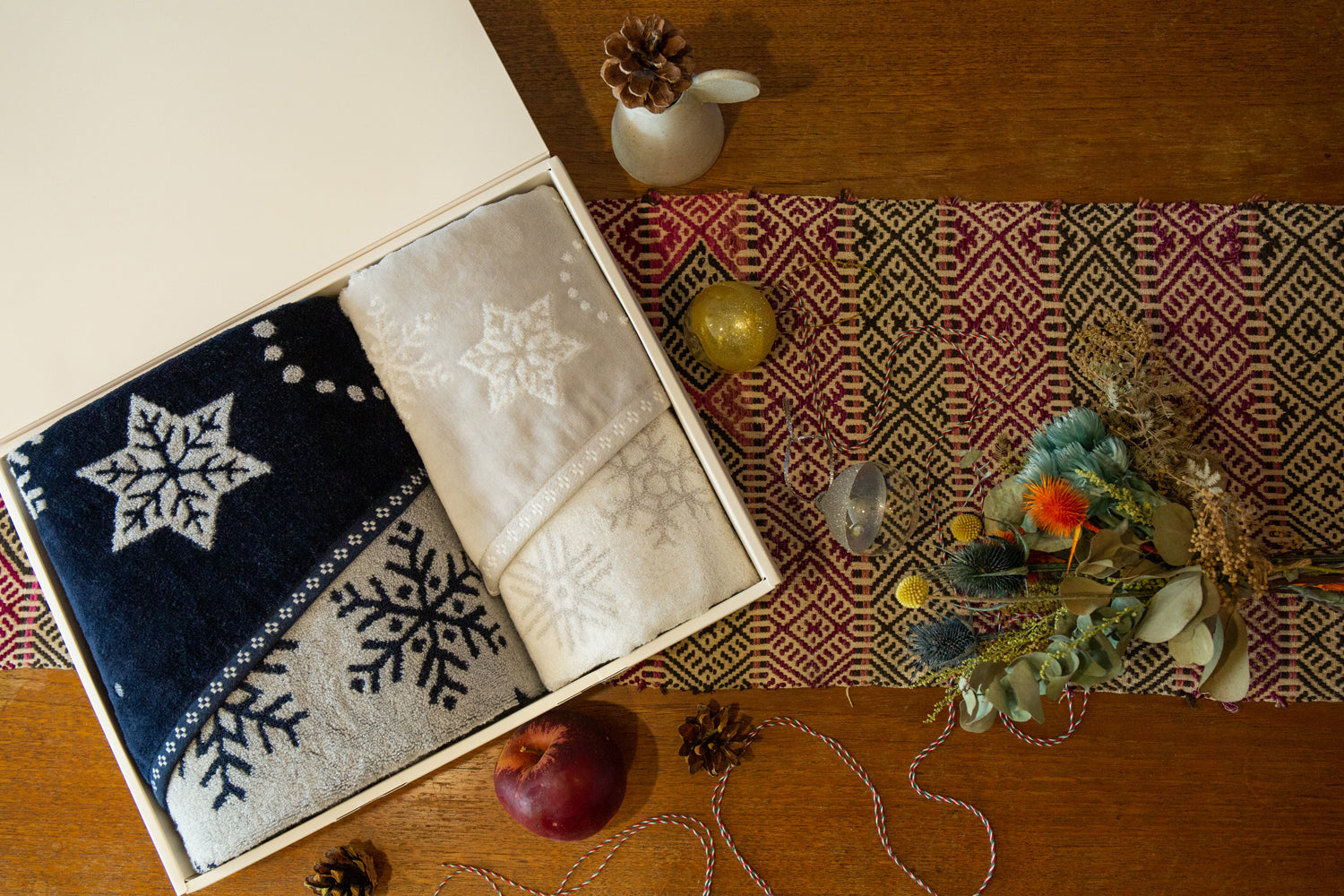 Two folded Imabari towels with snowflake patterns in a box on a wooden surface with decorative items.