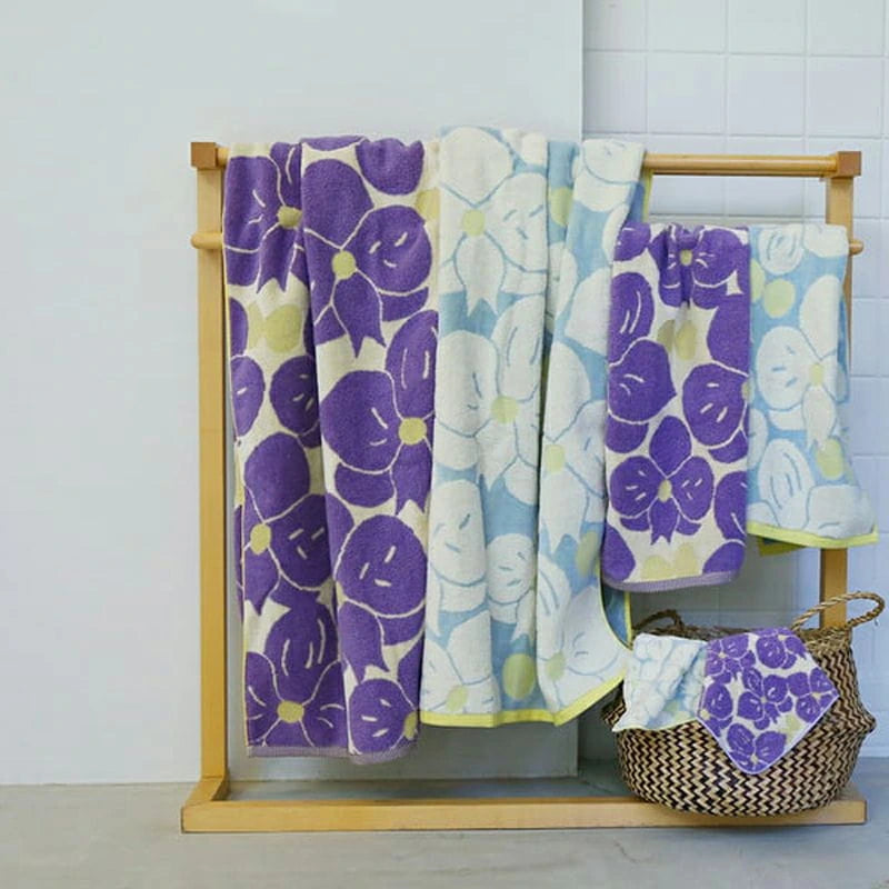 A set of Imabari bath towels with a jacquard weave featuring a phalaenopsis orchid motif in white and purple, displayed on a wooden rack and in a basket.
