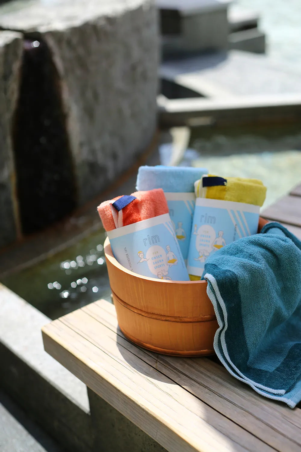Three colors of folded Imabari towels with paper bands, placed inside a wooden bath bucket