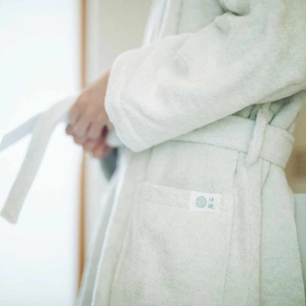 A close-up of a person wearing a soft, light cream-colored terry cloth Imabari towel bathrobe, with their hands adjusting the tie belt. A small woven brand tag is visible on the front pocket.