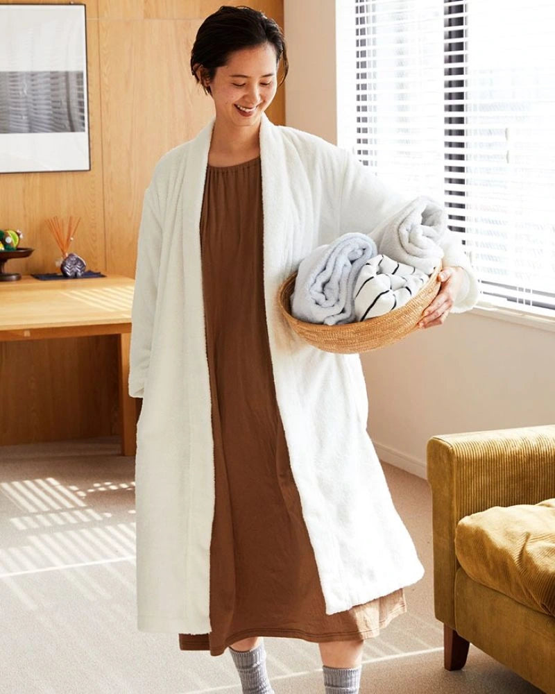 A woman in a brown dress wearing a long white Imabari towel bathrobe stands in a sunlit room, holding a woven basket with folded towels and blankets.
