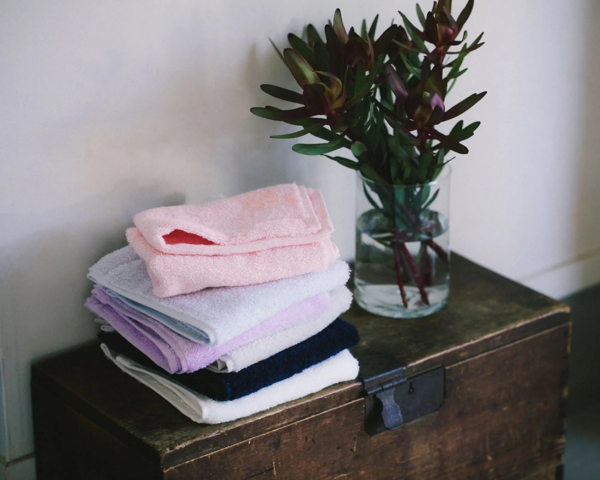 Stack of folded Imabari towels on a wooden chest with a vase of flowers.