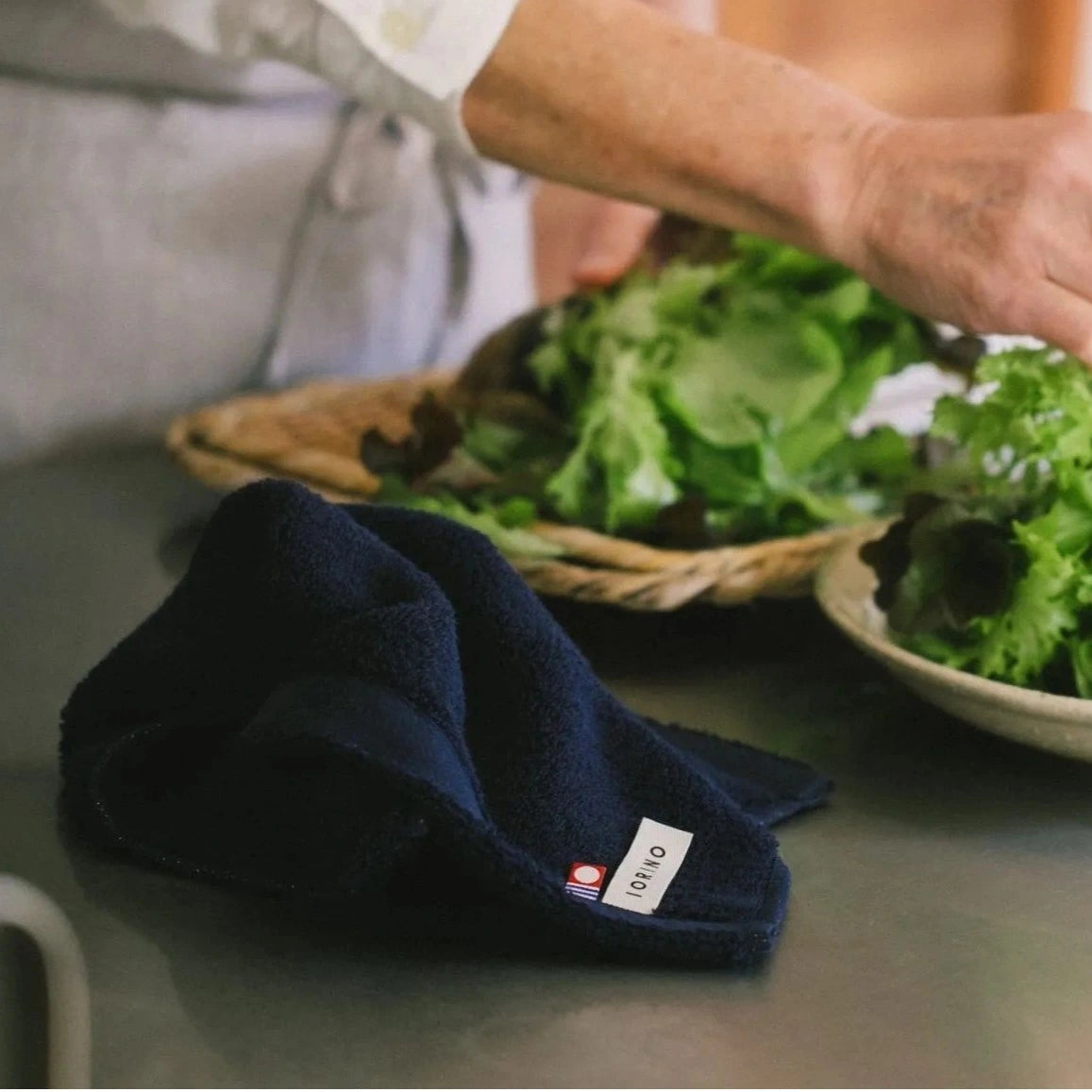 Navy blue Imabari towel with a brand label on a table next to a basket of green leafy vegetables.