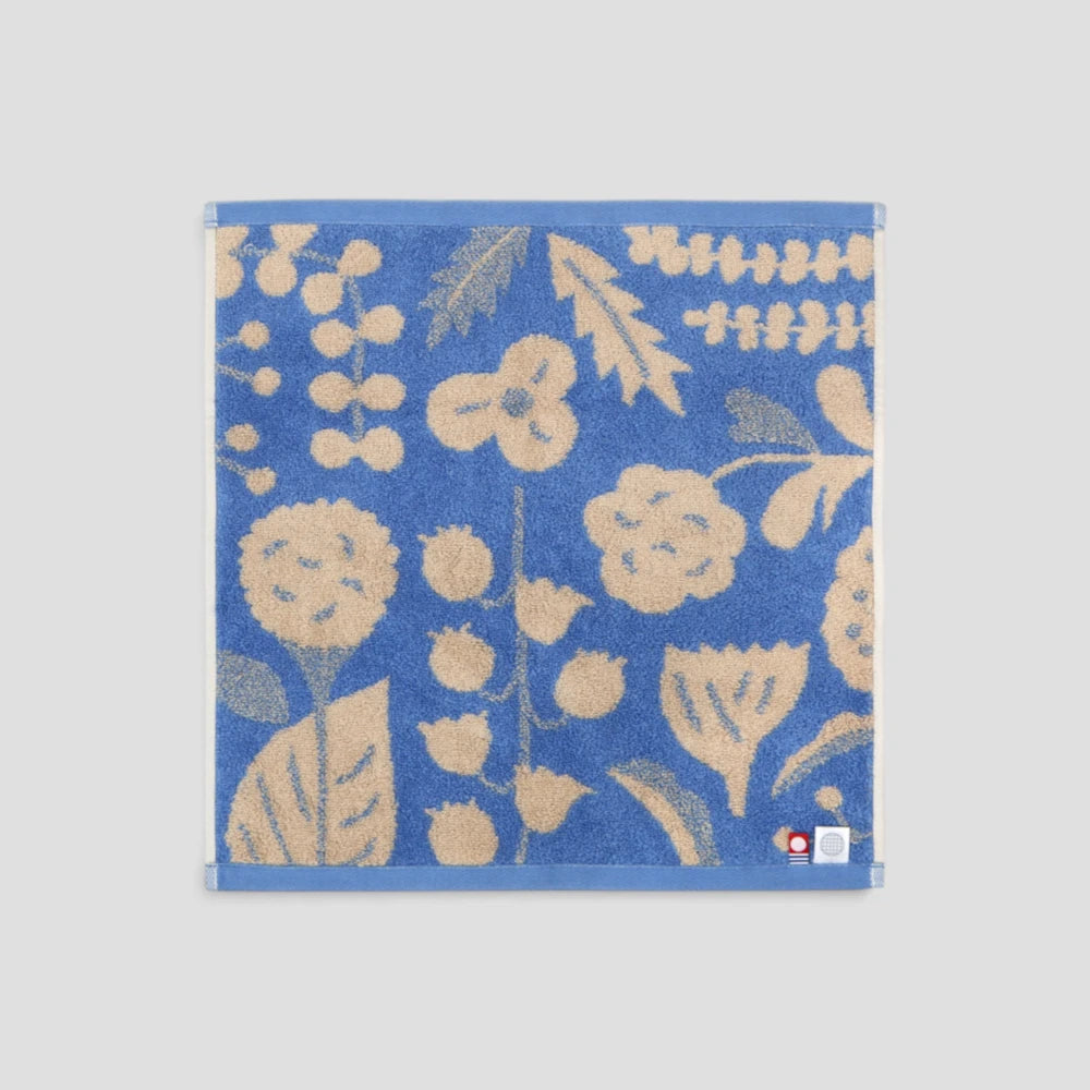 A square blue Imabari towel with a beige botanical pattern of flowers, leaves, and berries, displayed flat against a light gray background.