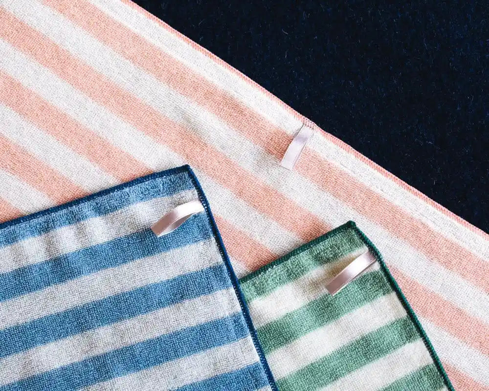 Two striped Imabari towels, one blue and white, one green and white, on a pink and white striped surface.