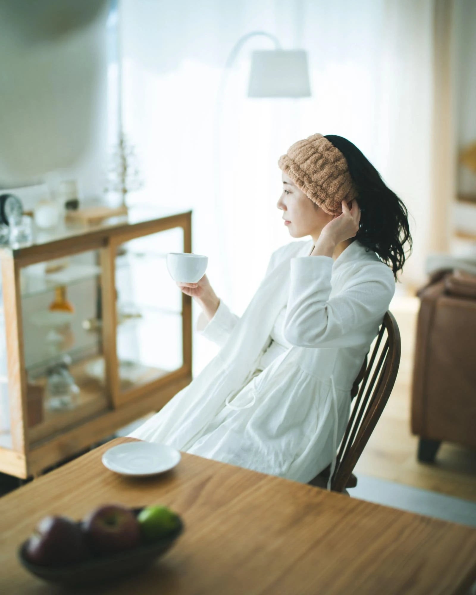 Woman sitting with beige Imabari towel head band at a table in a bright room, holding a cup.