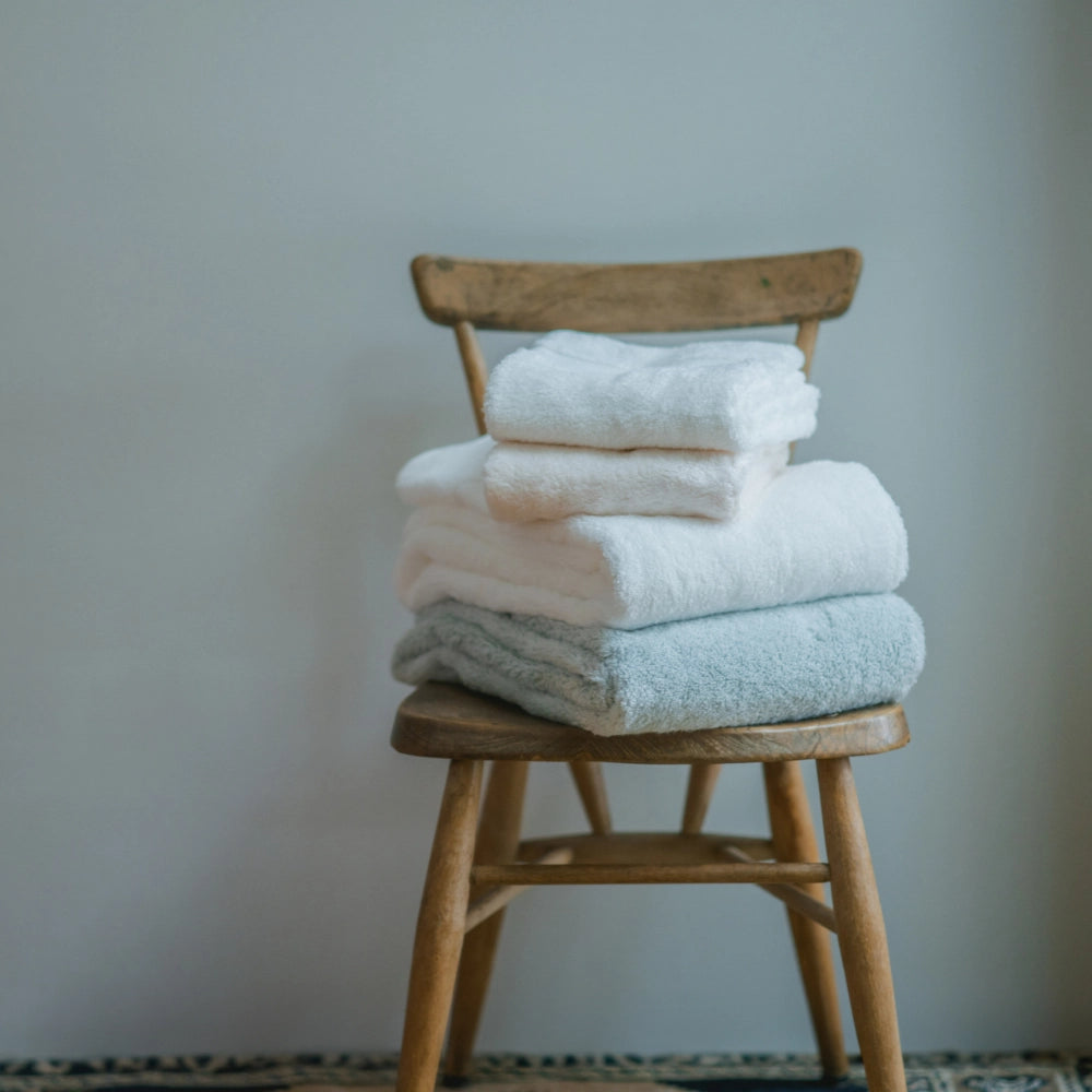 Stack of white and light blue Imabari towels on a wooden chair against a plain wall.