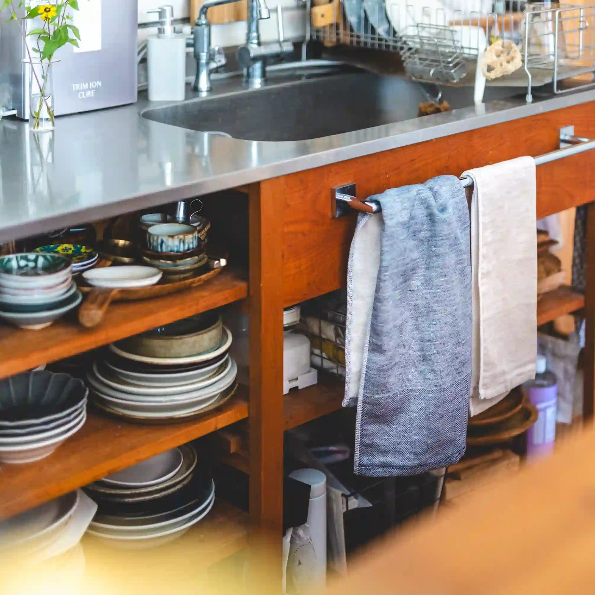 A warm, cozy kitchen scene featuring a wooden cabinet with a stainless steel countertop, a sink, and towels hanging from a towel bar. Open shelves below are stacked with plates and bowls, and a blurred wooden surface is in the foreground.