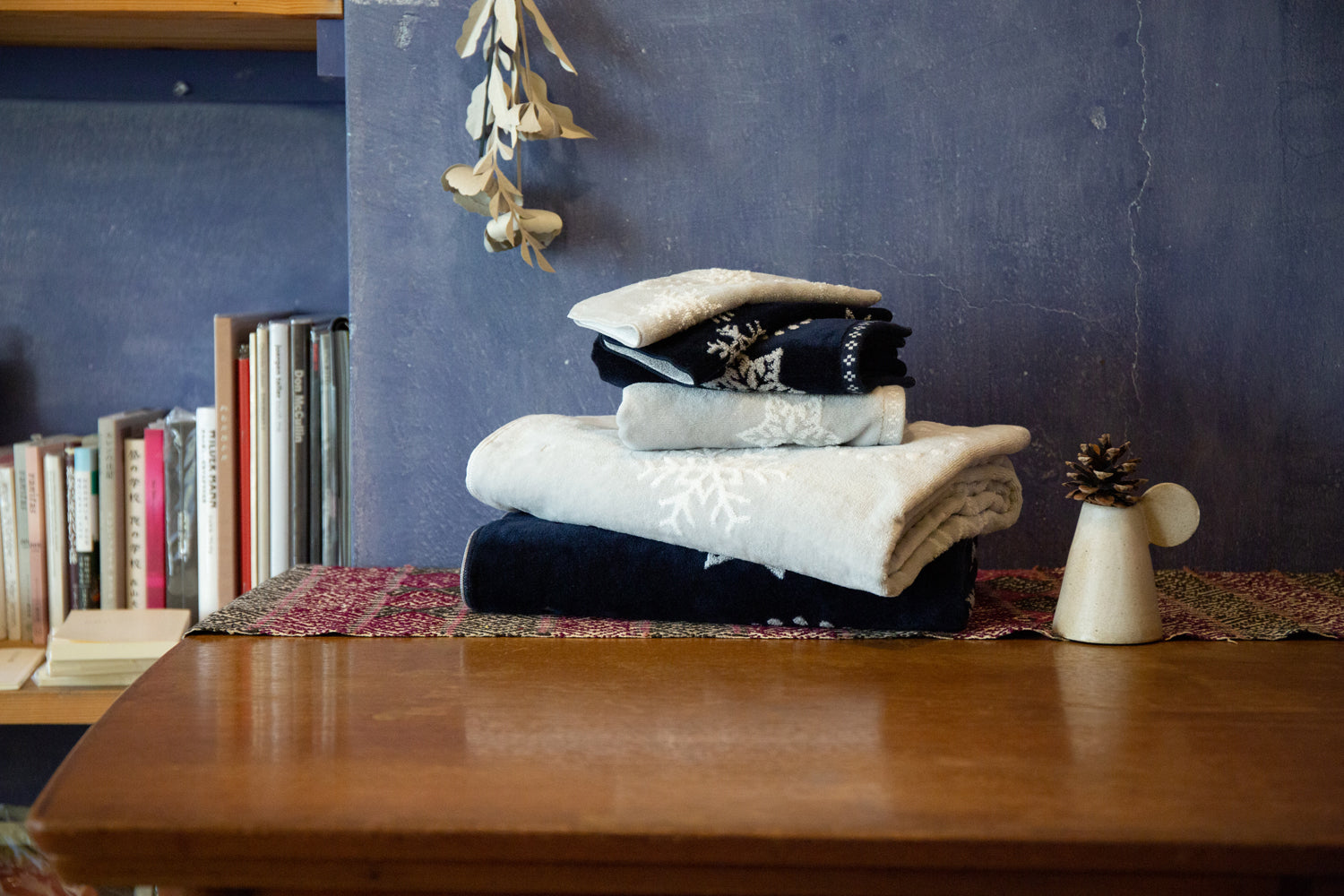 Stack of folded Imabari towels with a snowflake patterns on a wooden surface with books and decorative items in the background.