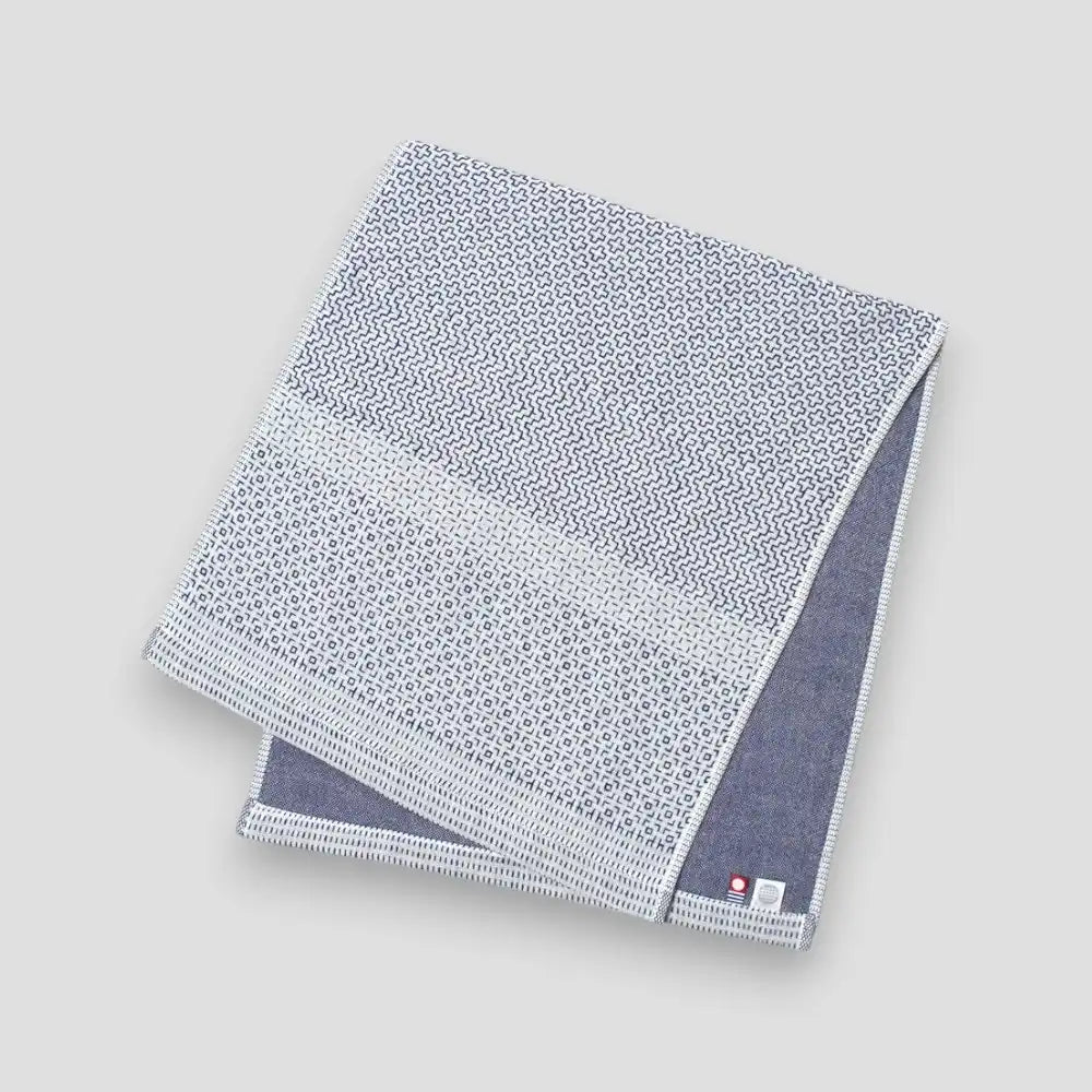 A folded white Imabari towel with a blue geometric pattern on one side, placed on a plain background.