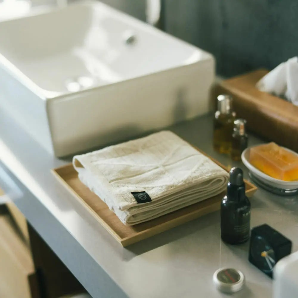 A folded white Imabari towel placed on a wooden tray near a sink.