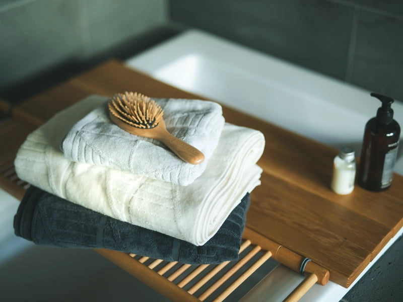 A set of folded Imabari towels in off-white and gray, with a wooden brush on top, placed on a wooden rack near a sink.