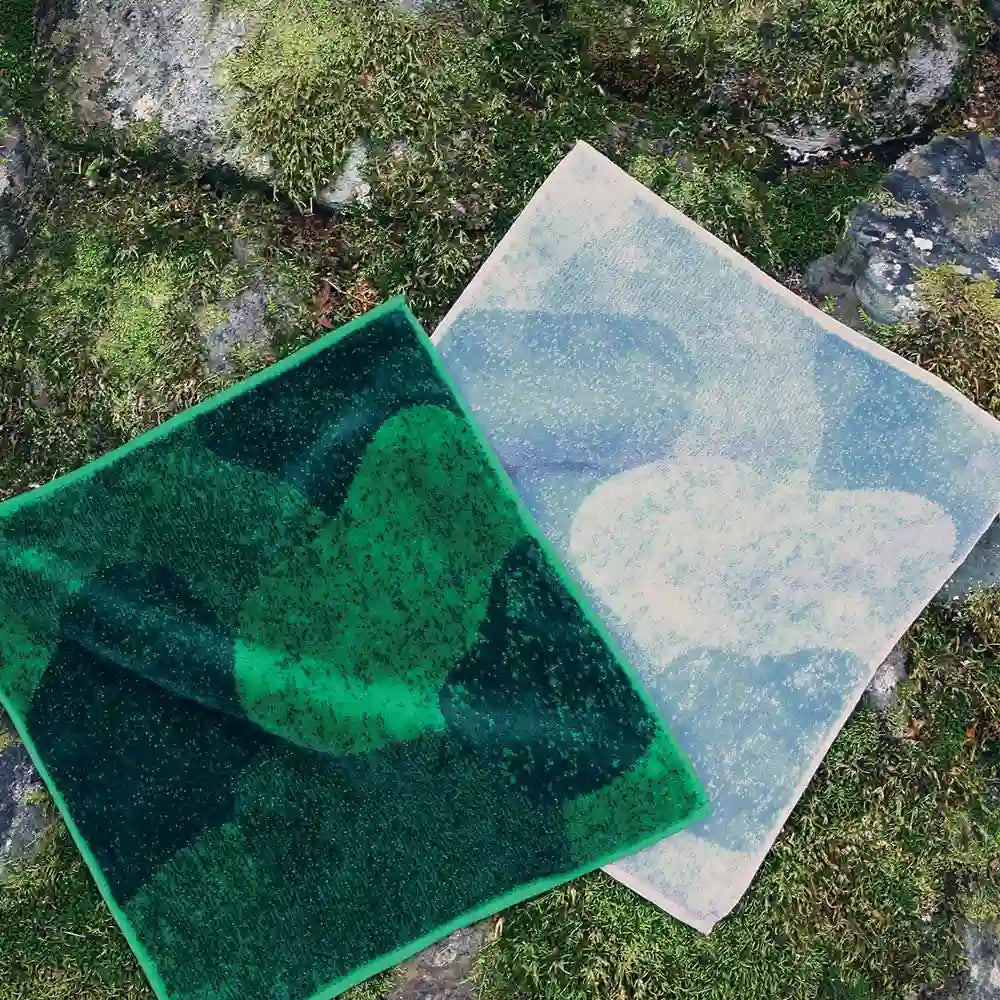 Two Imabari towels, one in green resembling moss and the other in gray resembling stone, laid out flat on the stone with moss