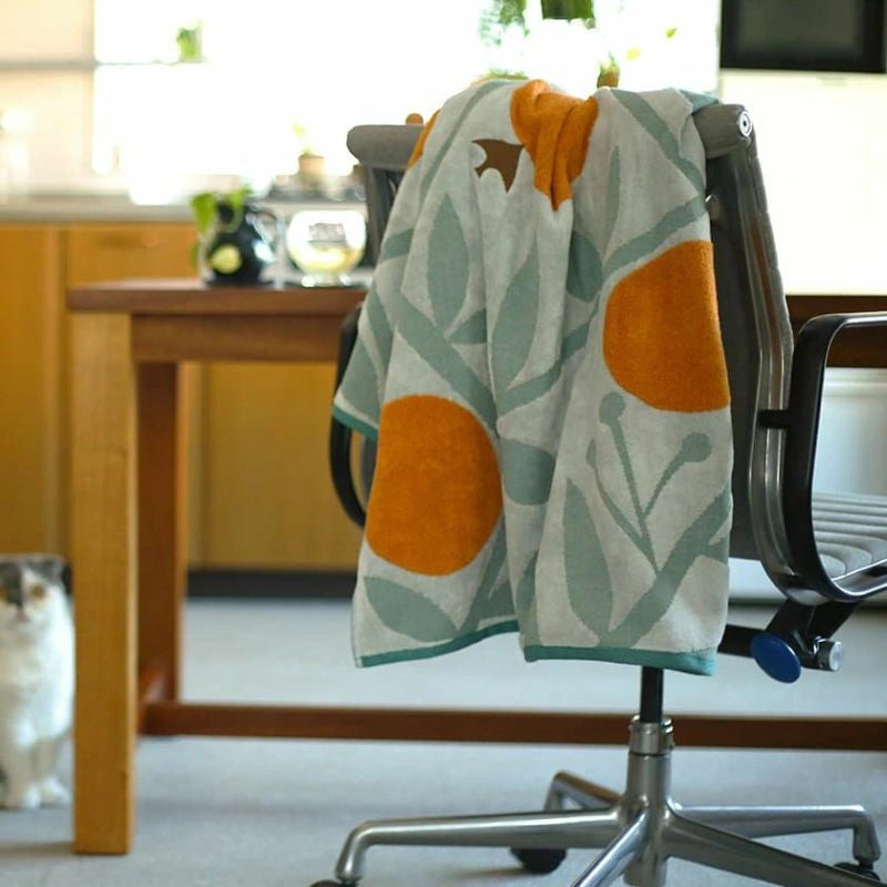 A multicolored Imabari towel with a bird pattern is hanging on a modern office chair, with a cat sitting on the floor in the background.