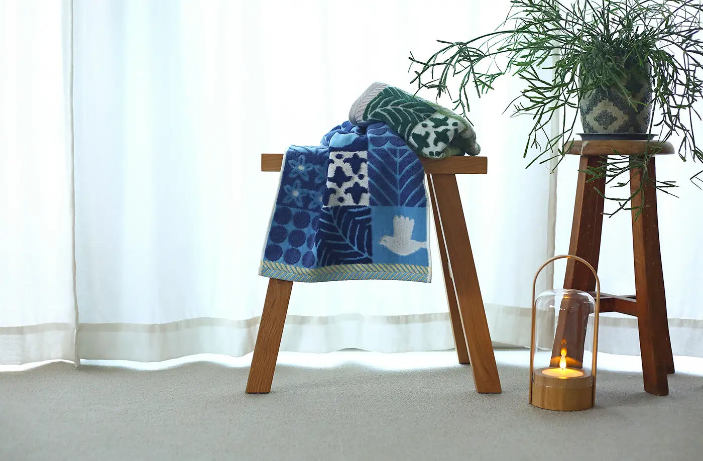 A blue and green patterned Imabari towel displayed on a wooden stool with a plant in the background and a candle holder with a lit candle in front.