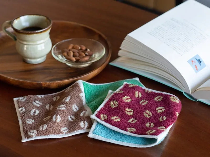 Two  compact Imabari towels in vibrant patterns leave on the table beside a cup of coffee and coffee beans.