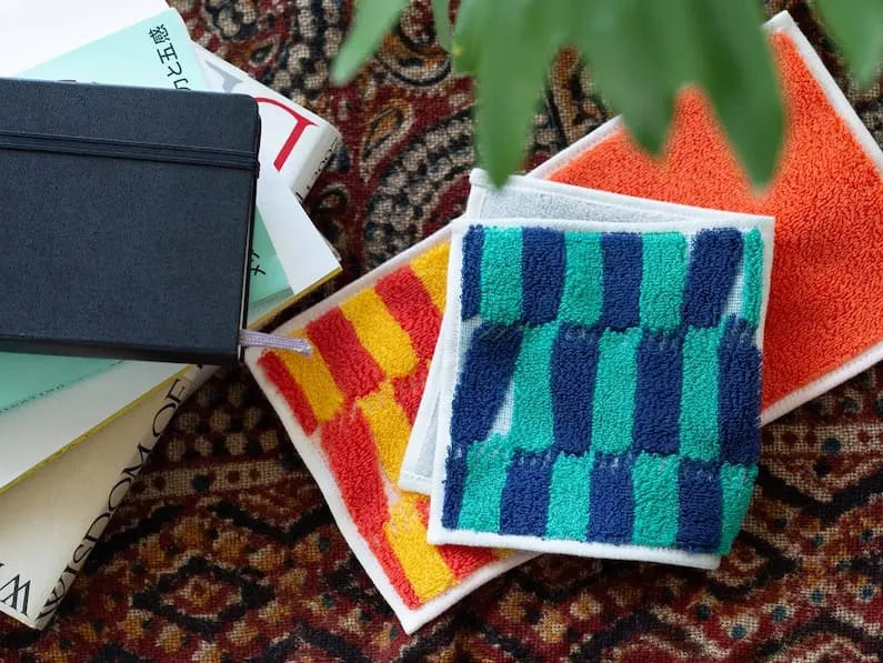 Two  compact Imabari towels in vibrant patterns leave on the desk beside some books.