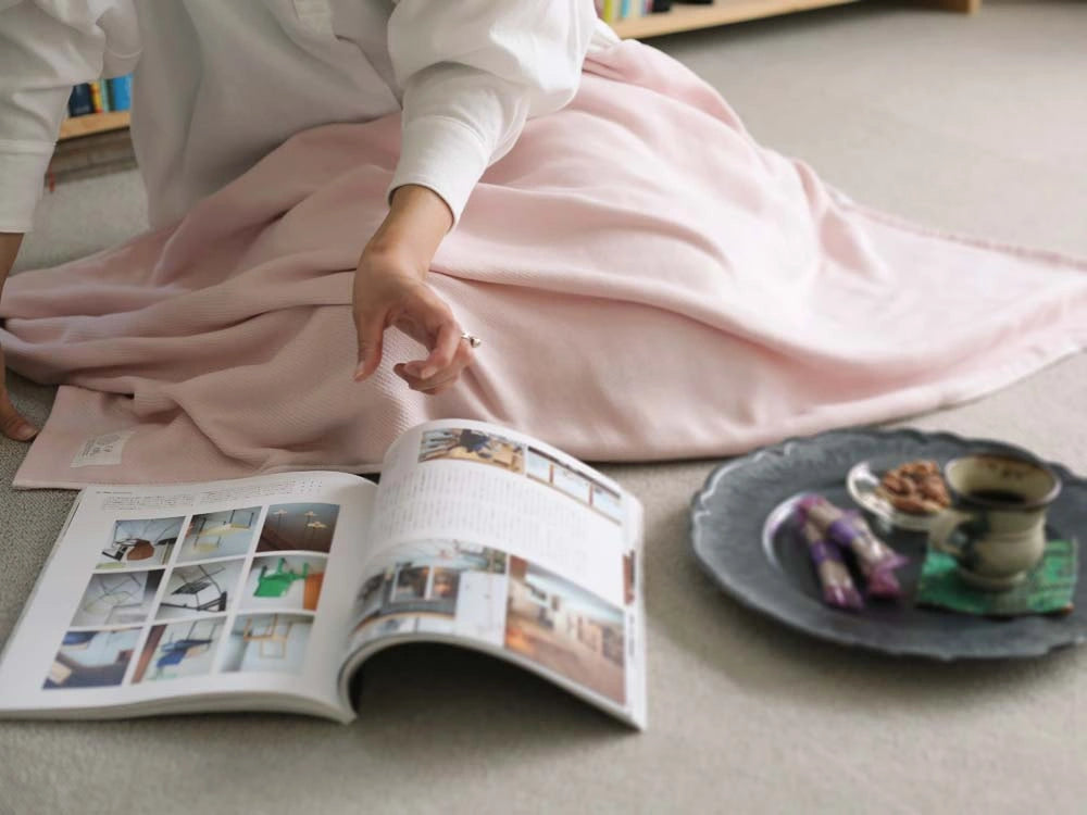 A woman sitting with an Imabari pink gauze towel wrapped around her knees, reading a magazine.