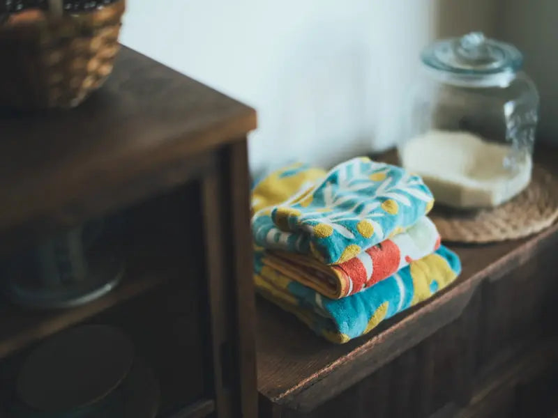 Stack of colorful Imabari towel on a wooden surface with a shelf of dishes in the background.