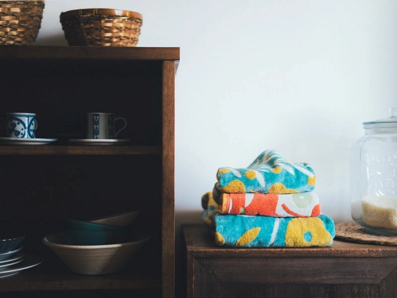 Stack of colorful Imabari towel on a wooden surface with a shelf of dishes in the background.