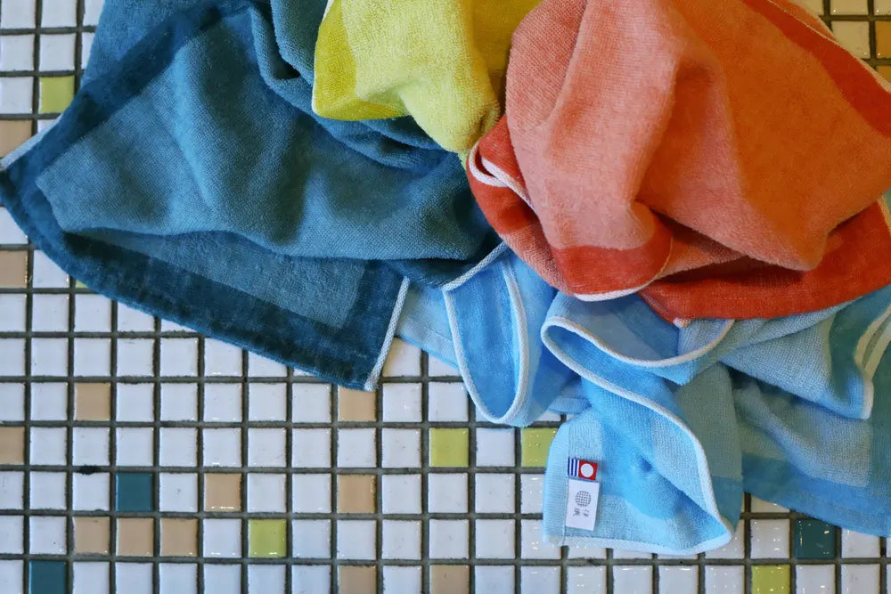 A collection of multi-purpose towels in various solid colors, including yellow, blue, green, and orange, placed on a tiled bathroom vanity