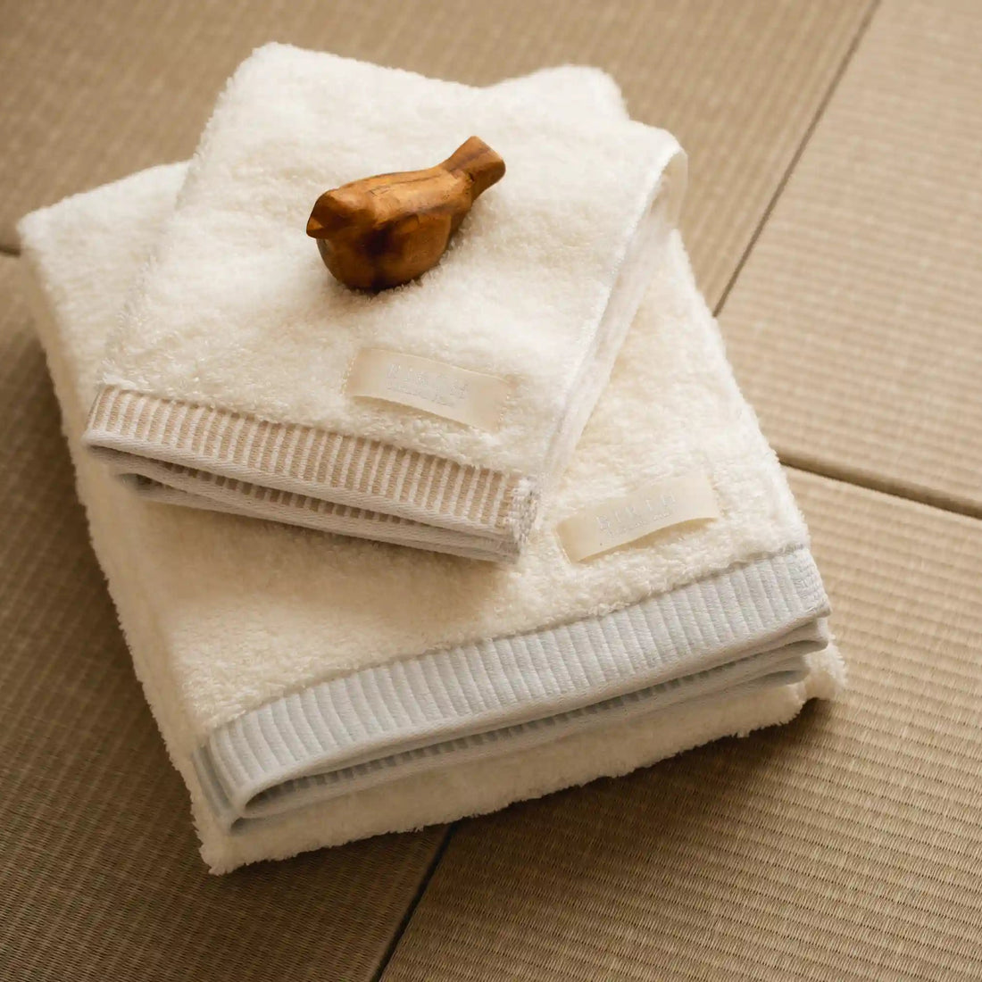 Two folded white towels stacked on a beige textured surface, with a small wooden bird-shaped object resting on the top towel.