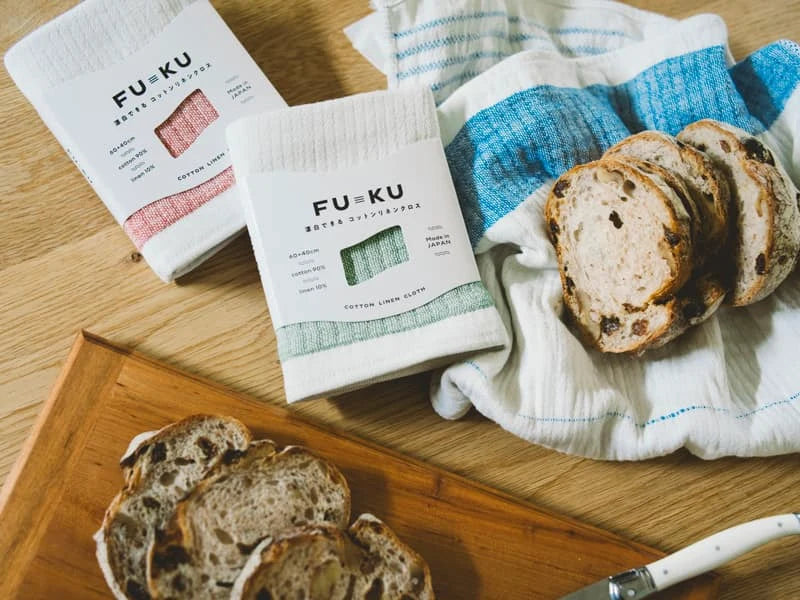 A set of FU-KU cotton Imabari towel  linen kitchen cloths in various colors displayed alongside sliced bread on a wooden table.