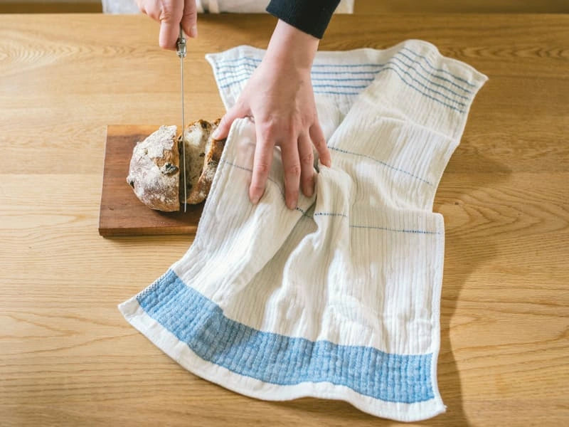 A woman slicing of bread holding Imabari towel linen kitchen cloth in blue line on left hand