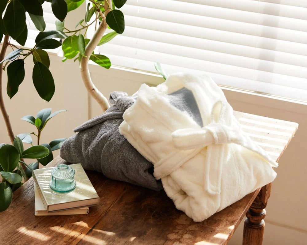 A plush cream Imabari towel bathrobe tied at the waist is draped over a folded gray imabari  towel bathrobe on a wooden table, with a window blind background, a potted plant, and a small stack of books with a glass candle holder nearby.