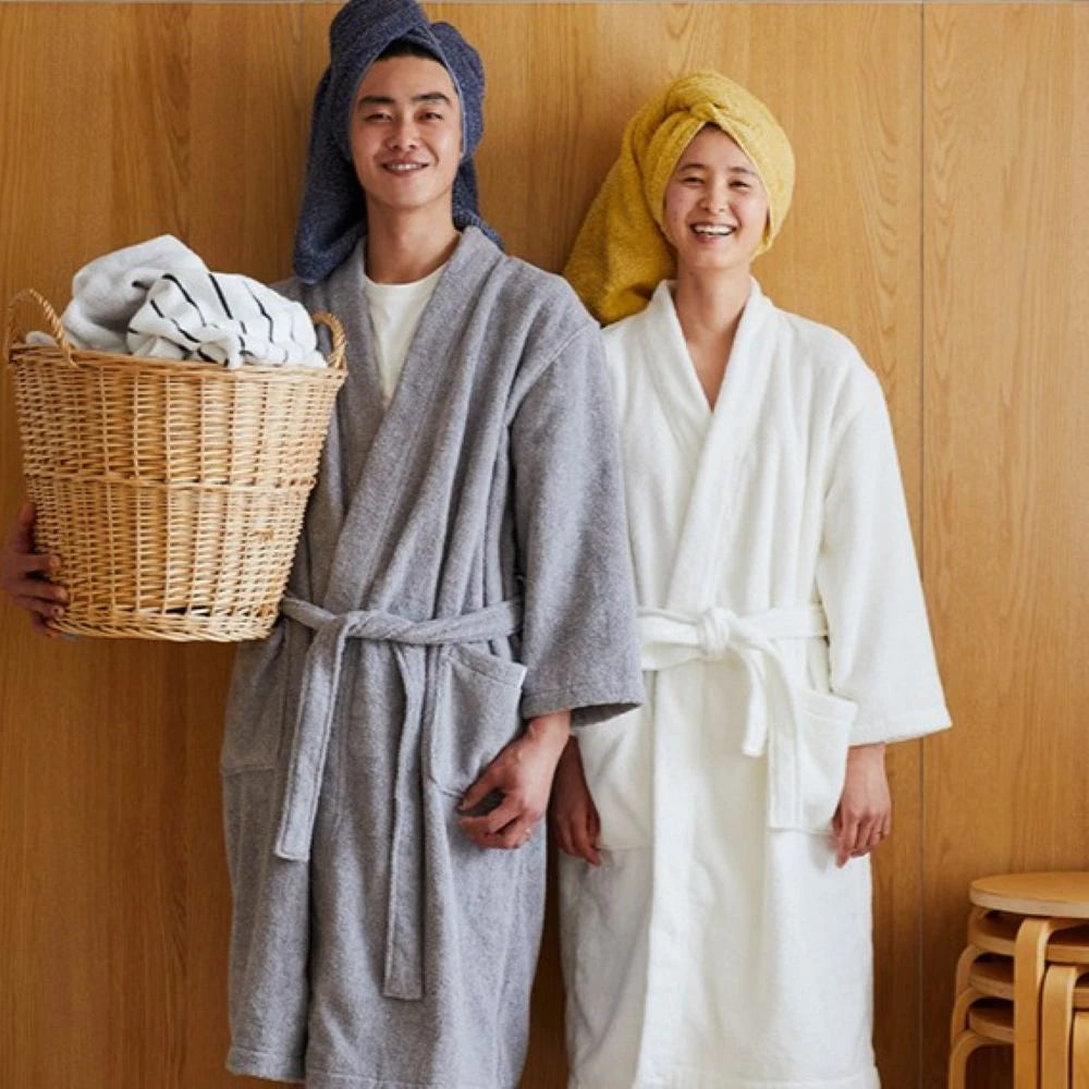 Two people standing indoors in front of a wood-paneled wall, each wearing a Imabari towel bathrobe and a towel wrap on their head. One person holds a wicker basket with folded white towels.