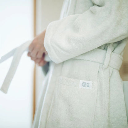 A close-up of a person wearing a soft, light cream-colored terry cloth Imabari towel bathrobe, with their hands adjusting the tie belt. A small woven brand tag is visible on the front pocket.