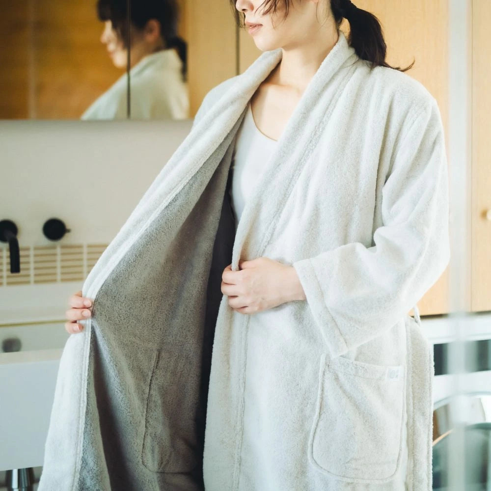 A woman wearing a plush white Imabari towel bathrobe with a gray lining, holding the robe open in a bright bathroom setting.