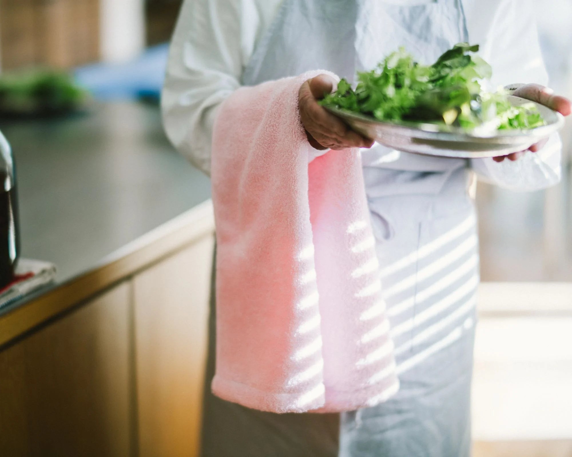 Person holding a plate of salad with a pink Imabari towel draped over their arm in a kitchen setting