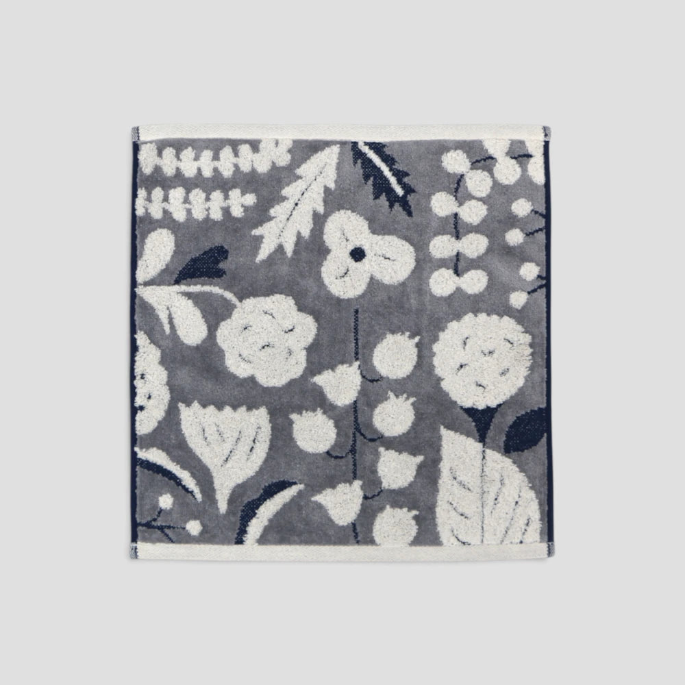 A square woven textile, likely a Imabari  hand towel or small rug, featuring a floral and botanical pattern in white and dark navy blue on a medium gray background, shown flat against a light gray backdrop.