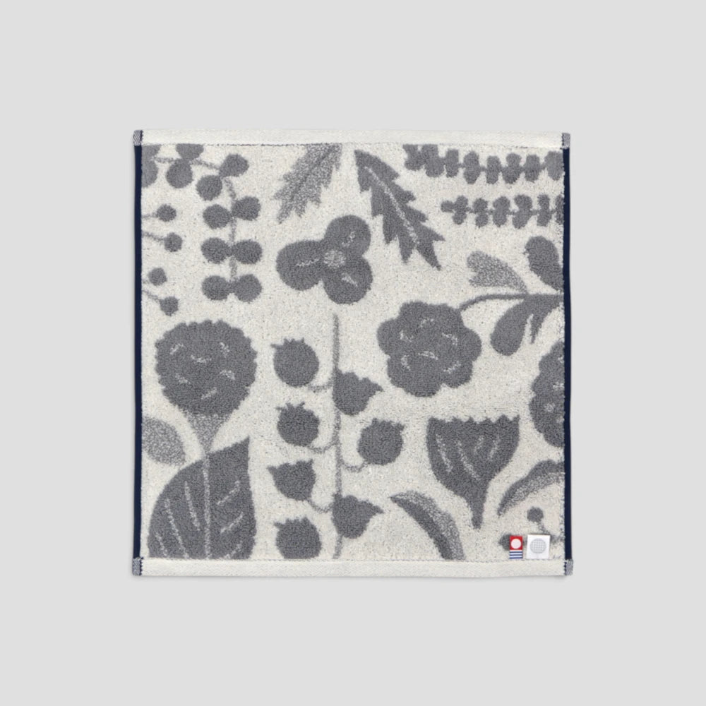 A square Imabari towel with a dark gray botanical and floral pattern on a light cream background, finished with a white border and a small red and white label in the bottom right corner, shown flat against a solid gray background.