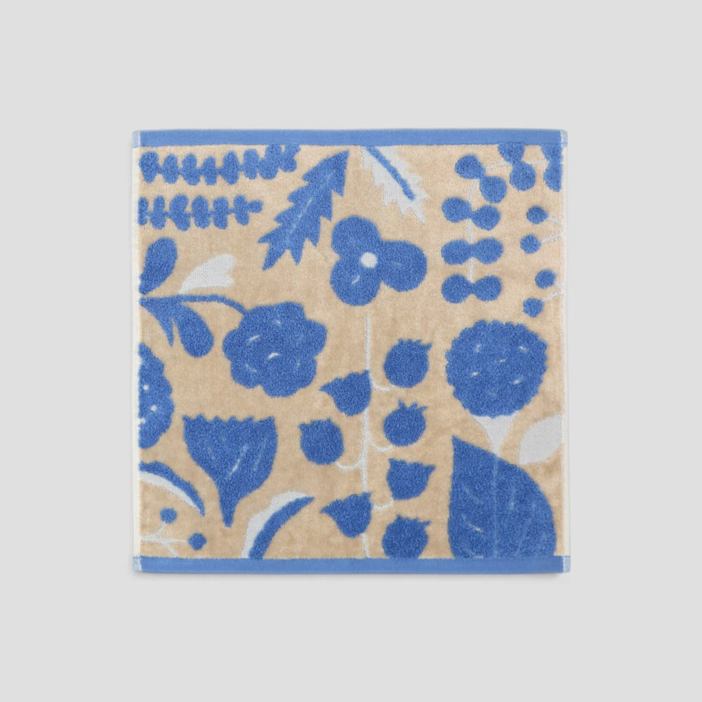 A square  small Imabari towel featuring a blue floral and botanical pattern on a warm beige background, with solid blue borders on two sides, shown against a light gray background.