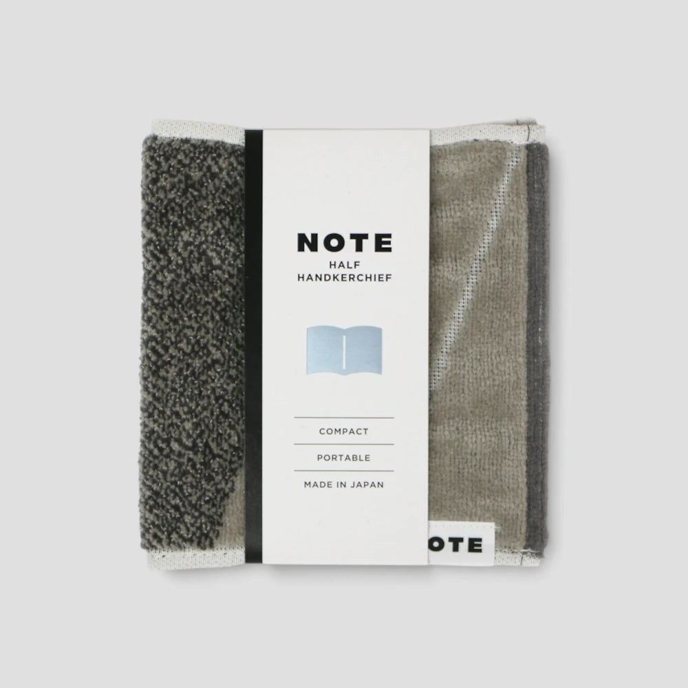 A packaged set of two compact Imabari towel handkerchiefs from the brand NOTE, shown on a light gray background. The left handkerchief is dark gray with a heathered texture, and the right one is a muted taupe-gray.