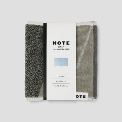 A packaged set of two compact Imabari towel handkerchiefs from the brand NOTE, shown on a light gray background. The left handkerchief is dark gray with a heathered texture, and the right one is a muted taupe-gray.