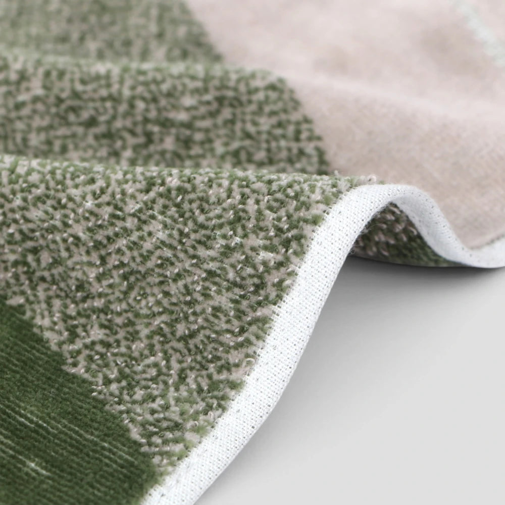 A close-up of a textured Imabari towel with a green and beige striped pattern, folded to show its plush looped texture and white stitched edge.