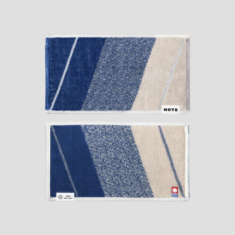 Two rectangular woven Imabari towels with a geometric pattern of navy blue, textured medium blue, and beige diagonal stripes, shown on a light gray background. The top towel has a white border and a small white &