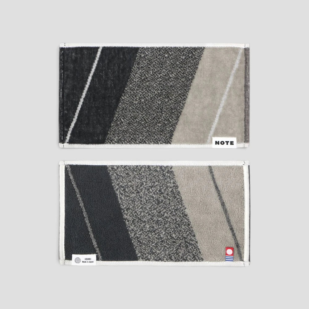 Two rectangular woven Imabari towels with a diagonal striped pattern in black, gray, and beige, displayed vertically on a light gray background. The top towel has a white border and a small white &