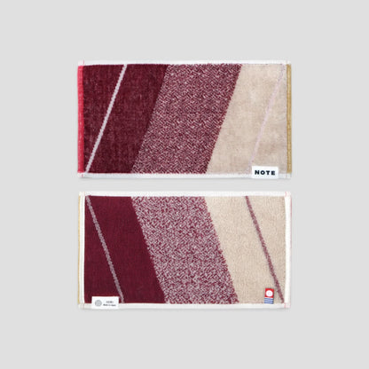Two rectangular Imabari towel with a geometric diagonal stripe pattern in burgundy, mauve, and beige, displayed vertically on a light gray background. The top towel has a white label reading &
