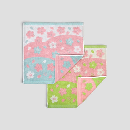 Two folded cotton Imabari towel  with a Japanese cherry blossom pattern, shown on a neutral light gray background. The Imabari towels feature pink, light blue, and light green sections with white and pink floral motifs.