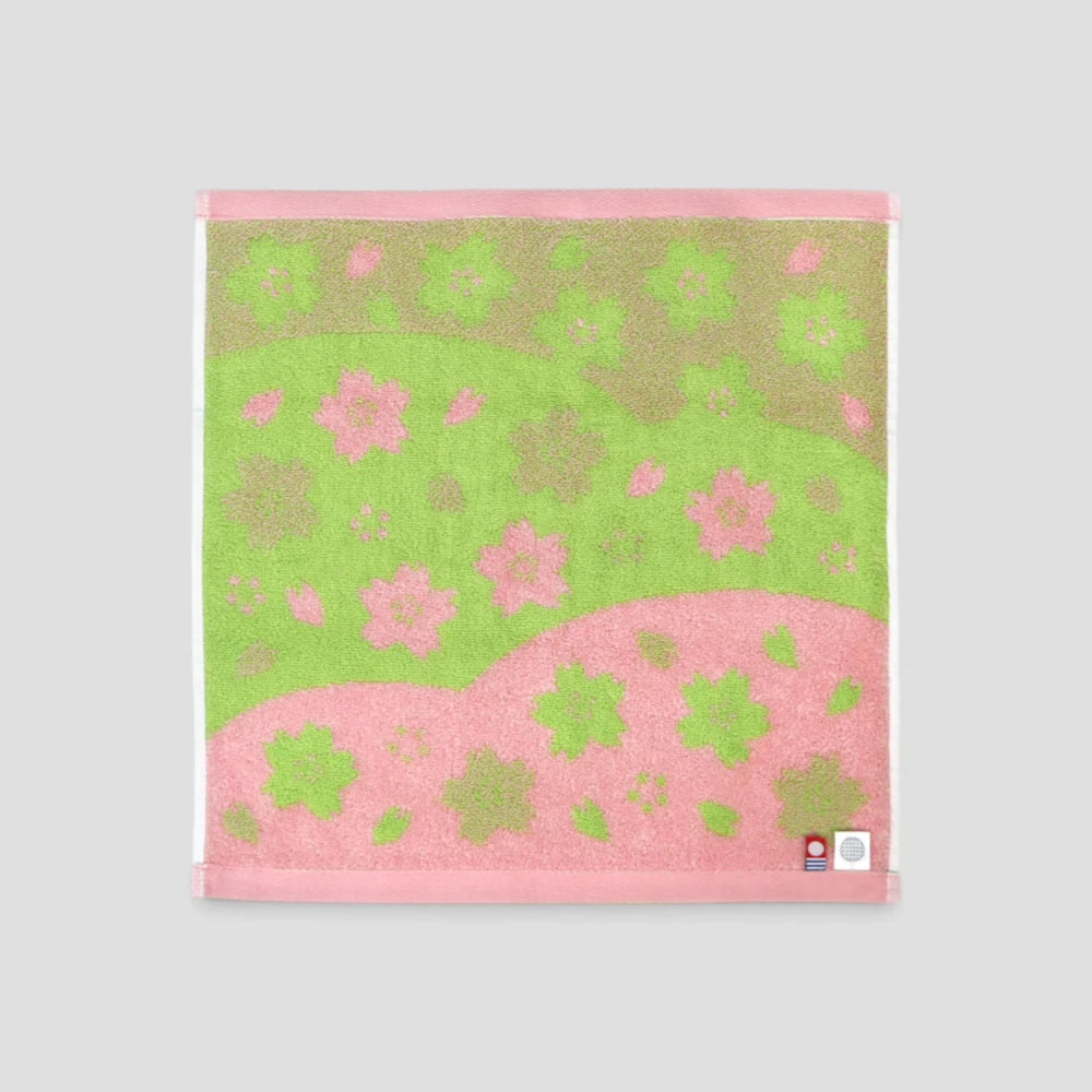 A square Imabari wash towel with a pink and green floral pattern, featuring a textured design with a pink border and a small Japanese tag in the bottom right corner, set against a solid gray background.