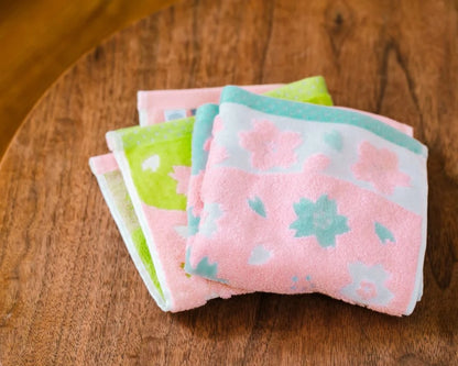 Three folded cotton Imabari towels with pink, green, and light blue floral patterns stacked on a round wooden table.