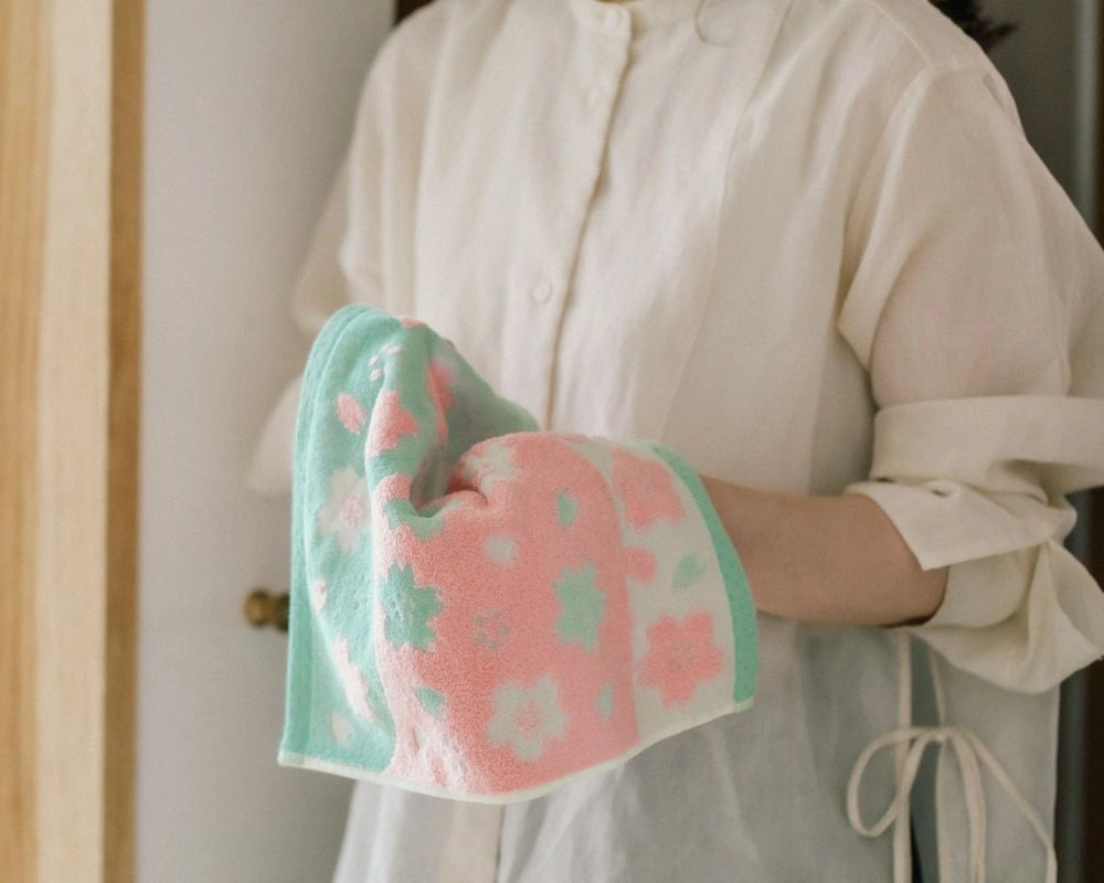 A person wearing a cream-colored linen shirt holds a soft, folded cotton Imabari towel with a pink and mint green floral pattern.