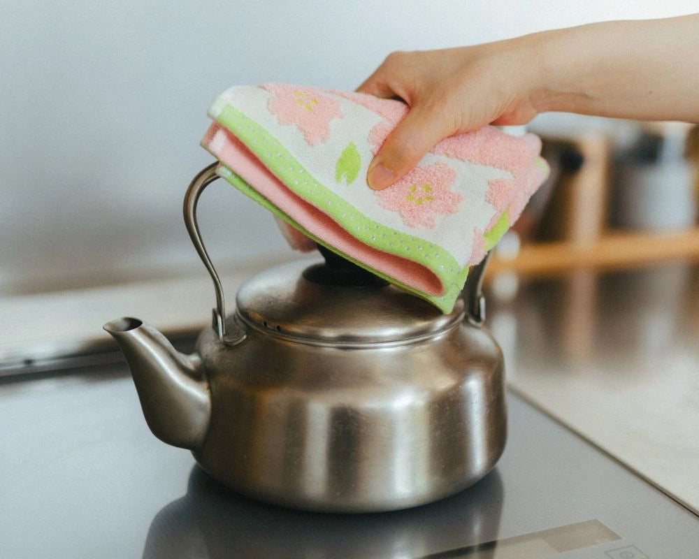 A hand holding a pink and green floral patterned Imabari towel while lifting the lid of a stainless steel kettle on a dark countertop.