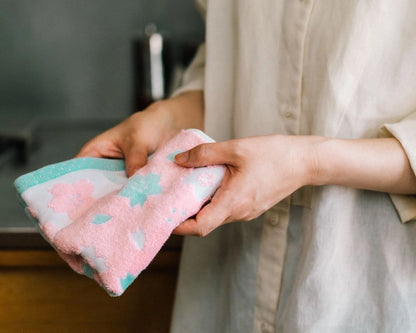 A person in a light beige linen shirt holds a folded pink cotton Imabari towel with a light blue and white patterned border in a kitchen setting.