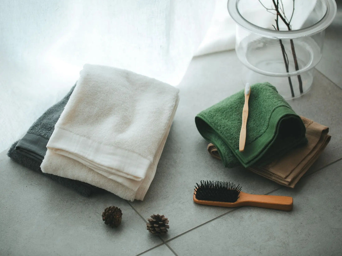 Folded Imabari towels, a brush, and pinecones on a tiled floor.
