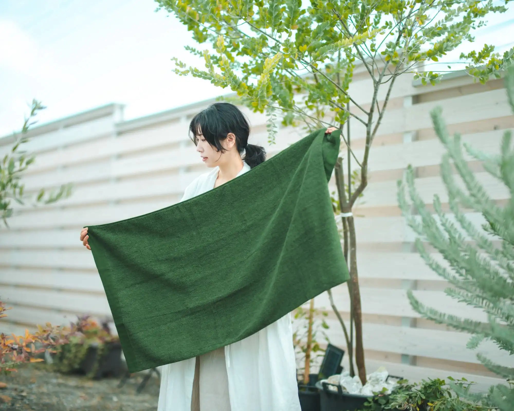 Woman holding a green Imabari towel in an outdoor setting with plants and a white wall.